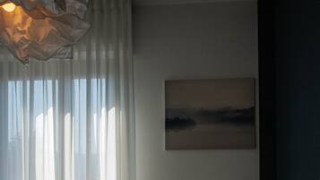 A bedroom with a bed, a radiator, a window with sheer curtains, and a painting on the wall.
