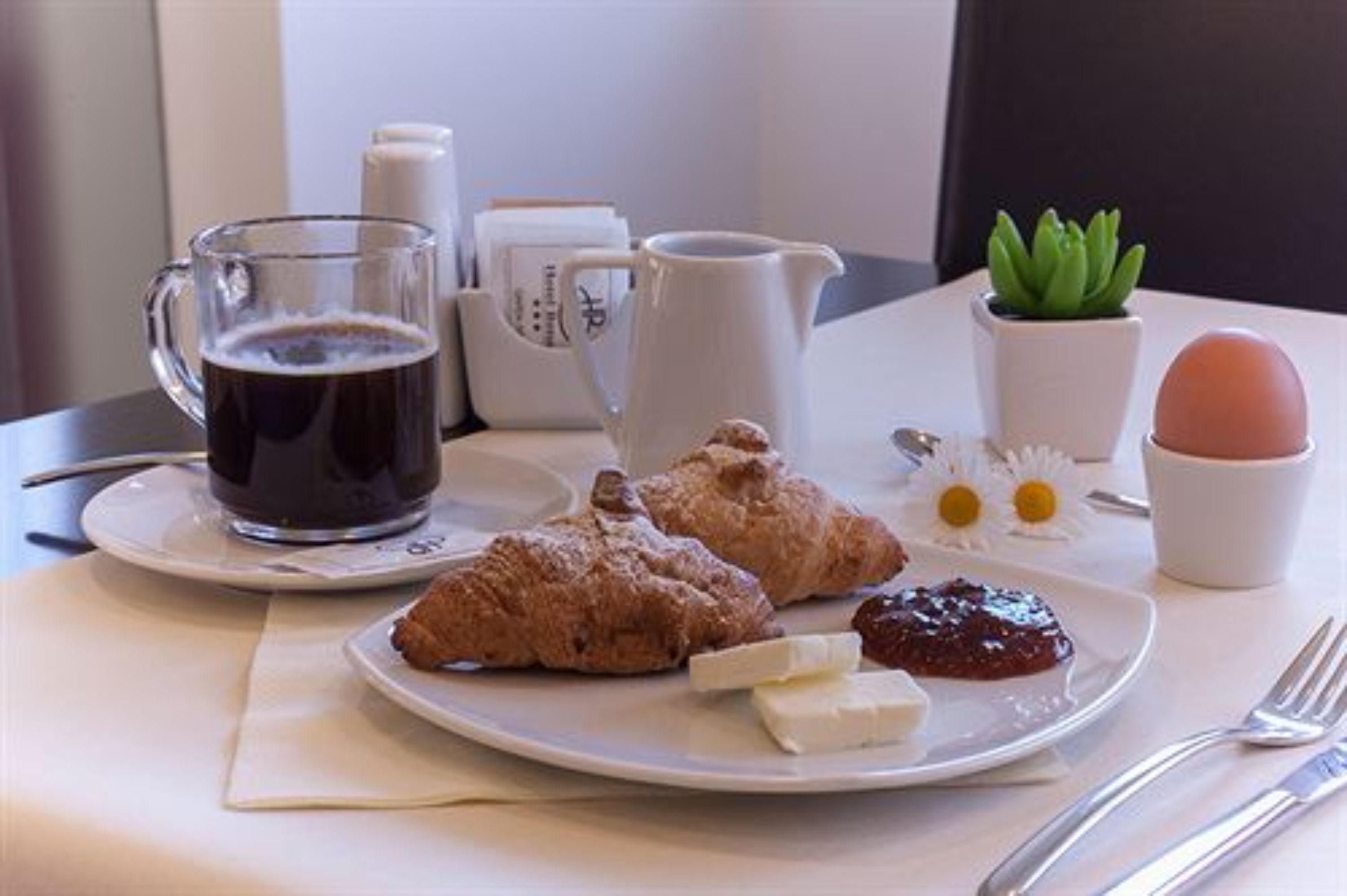 A breakfast setting with a coffee mug, croissants, a plate with jam and cheese, a pitcher, a small plant, and a cup with an egg.
