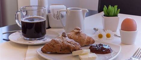 A breakfast setting with a coffee mug, croissants, a plate with jam and cheese, a pitcher, a small plant, and a cup with an egg.
