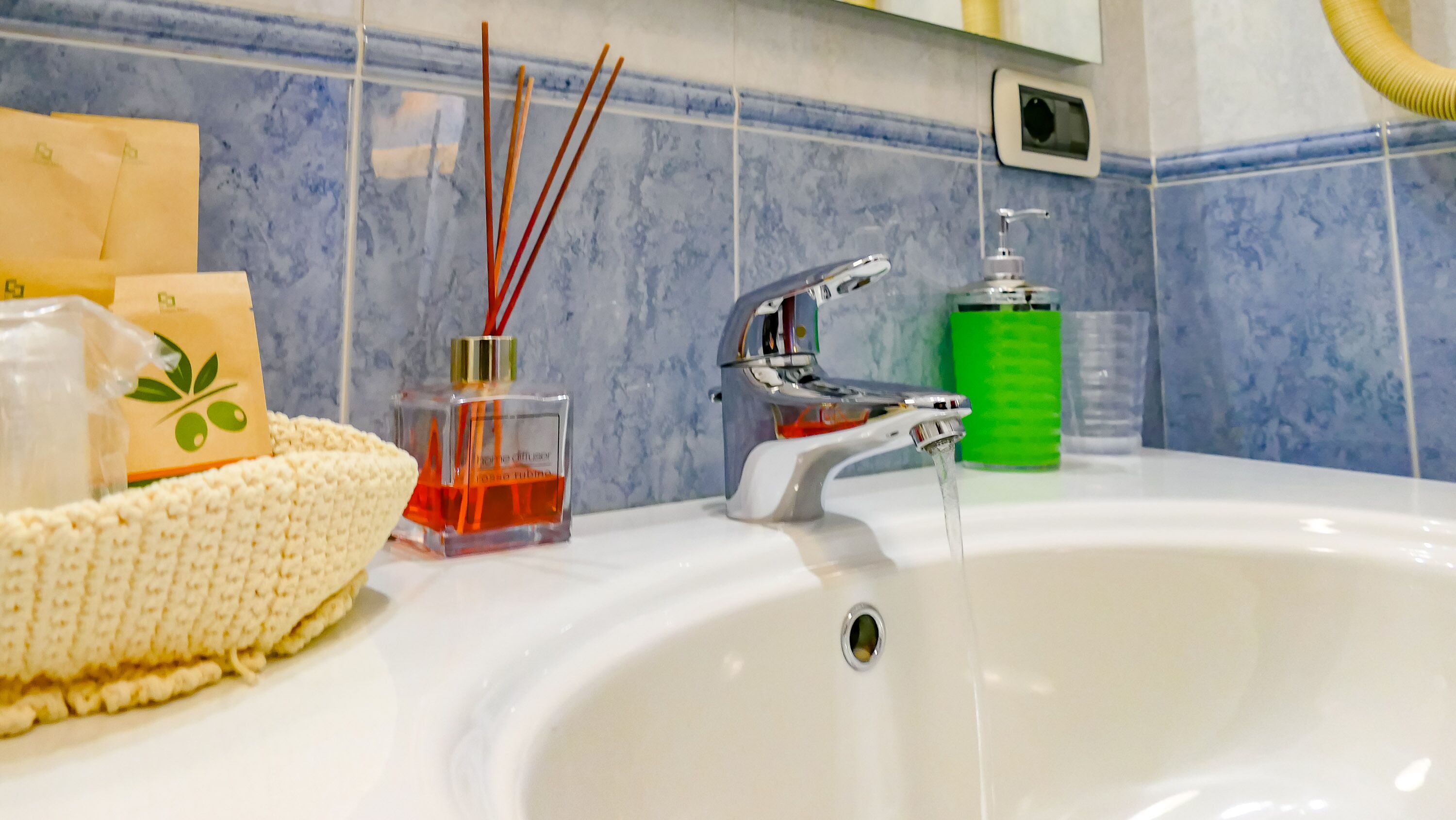 A bathroom sink with a chrome faucet, a green soap dispenser, and a small glass bottle with red reeds.