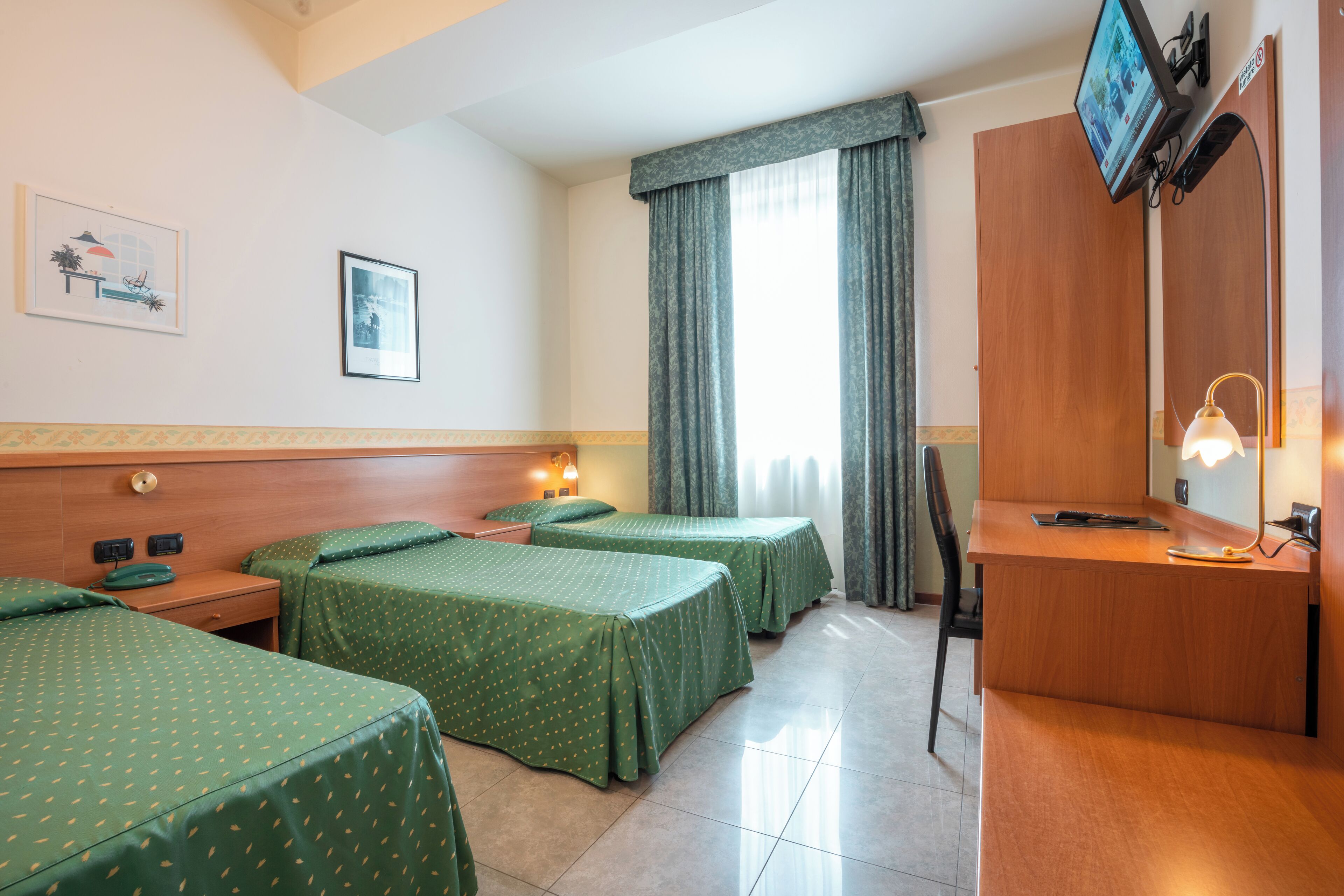 Habitación de hotel con dos camas individuales, un escritorio, una silla, un televisor y un ventanal con cortinas.