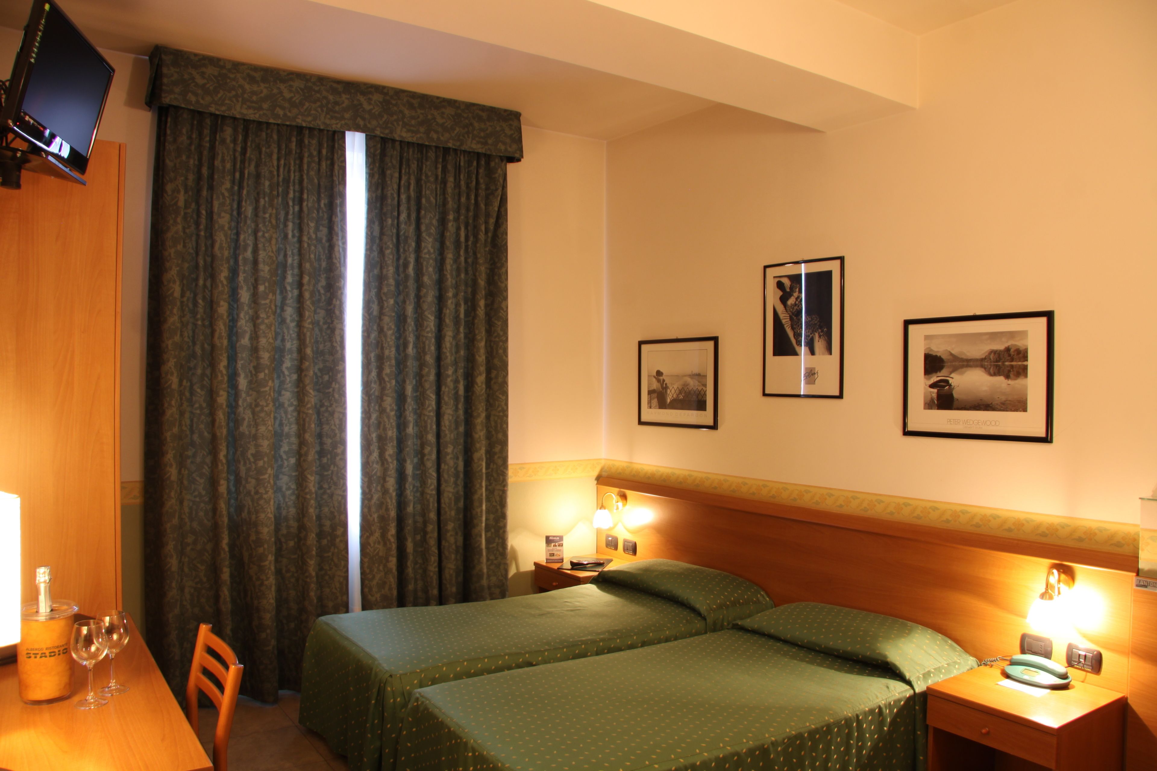 A hotel room with a bed, a desk with a bottle and glasses, a TV, and two framed pictures on the wall.