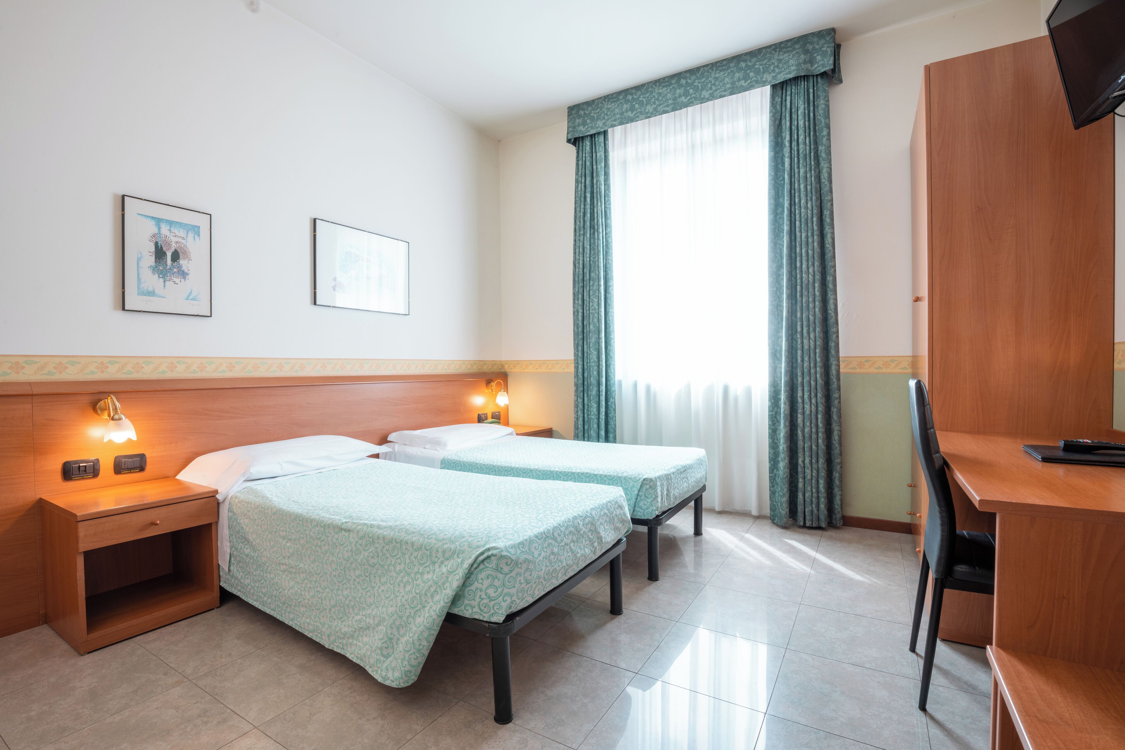 Una habitación de hotel con dos camas individuales, un escritorio, una silla, una mesita de noche con lámpara y una ventana con cortinas.
