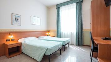 Una habitación de hotel con dos camas individuales, un escritorio, una silla, una mesita de noche con lámpara y una ventana con cortinas.