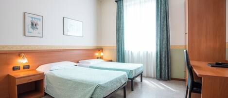 A hotel room with two single beds, a desk, a chair, a nightstand with a lamp, and a window with curtains.