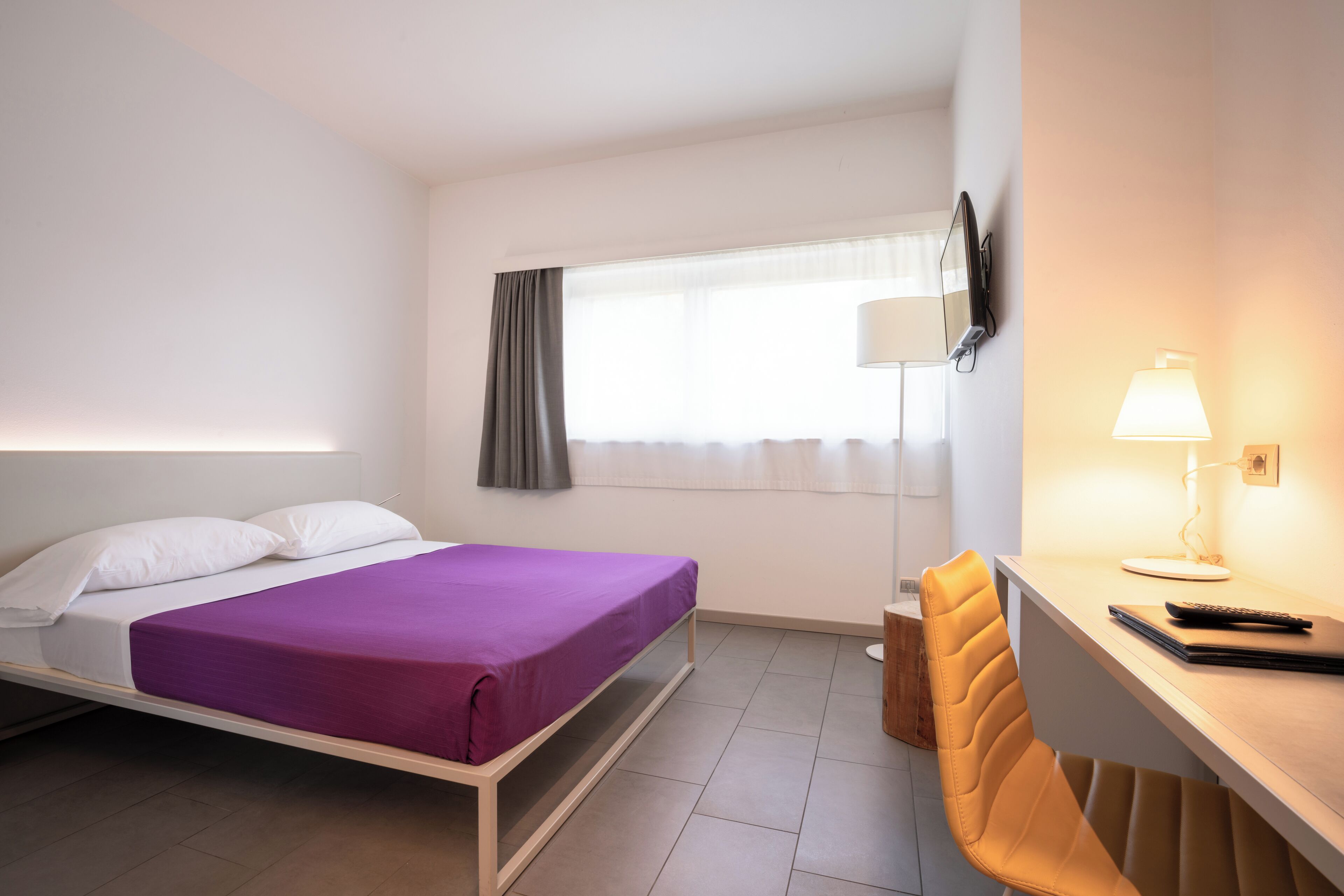 A hotel room with a bed, a desk, a chair, a lamp, and a window with curtains.