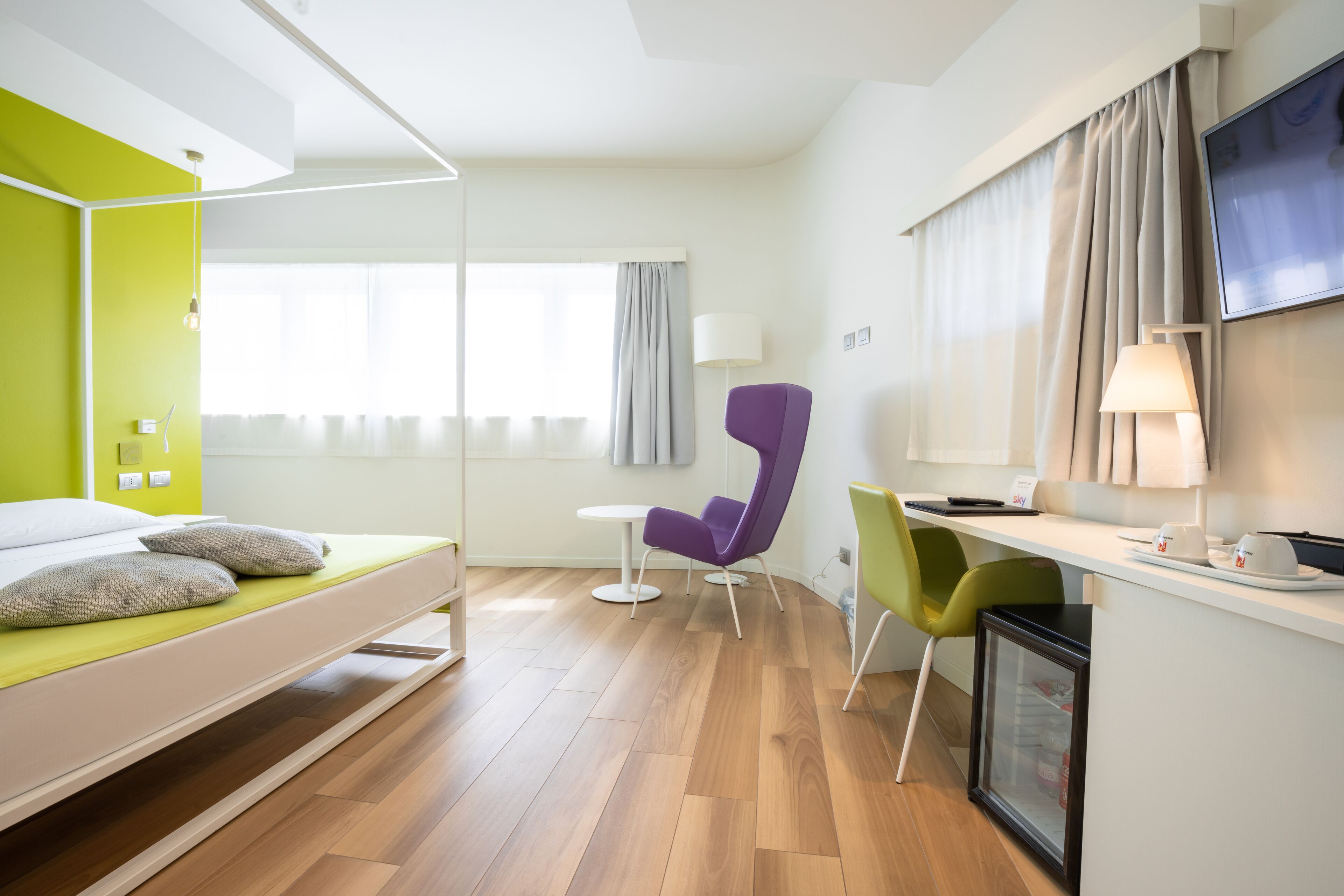 A modern hotel room with a bed, a desk, a chair, a TV, and a mini-fridge.