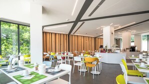 A modern dining area with white tables and chairs, a central counter, and large windows offering a view of greenery.
