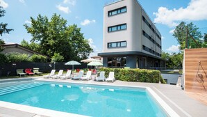 A swimming pool with lounge chairs and umbrellas, surrounded by greenery and a modern building.