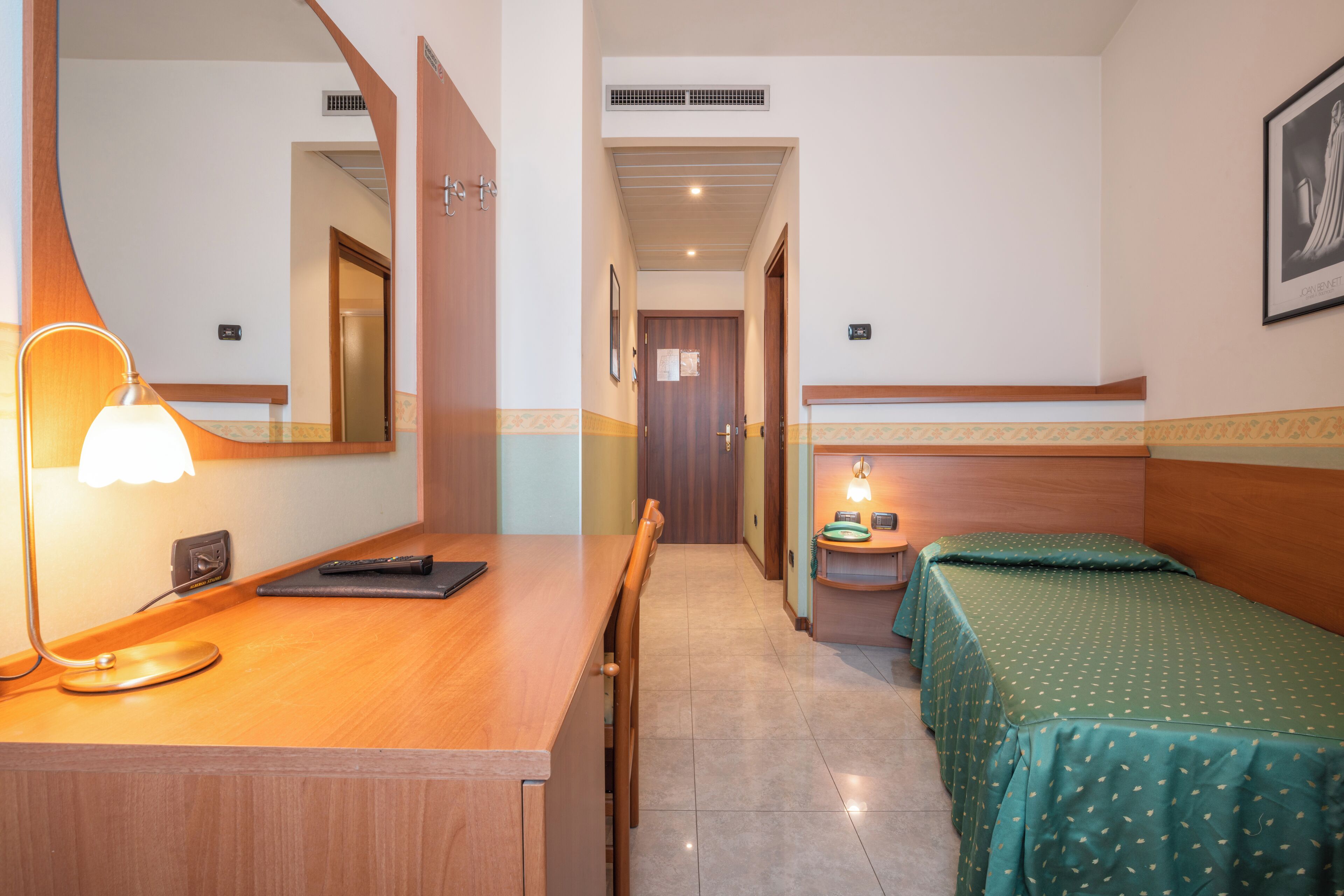 A hotel room with a wooden desk, a lamp, a bed with green bedding, a small bedside table with a lamp, and a mirror.
