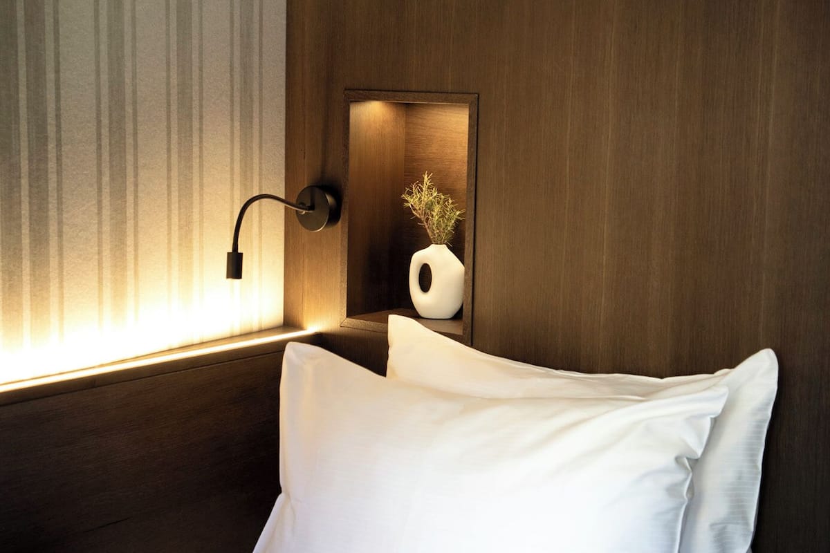 Una habitación de hotel con una cama, una lámpara de noche, un jarrón con flores secas y un ventanal con cortinas.