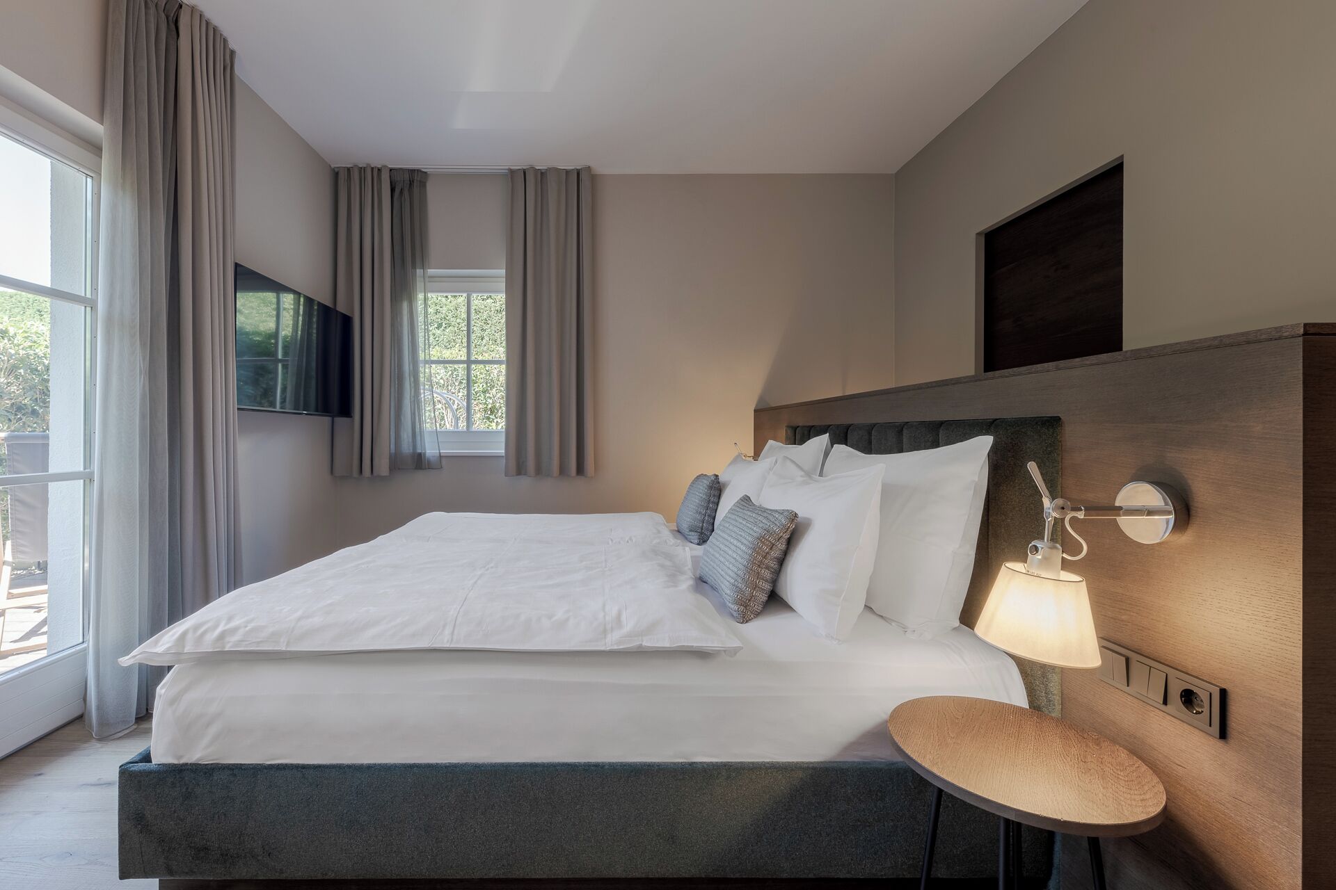 A neatly made bed with white linens and pillows, a bedside table with a lamp, and a window with curtains.