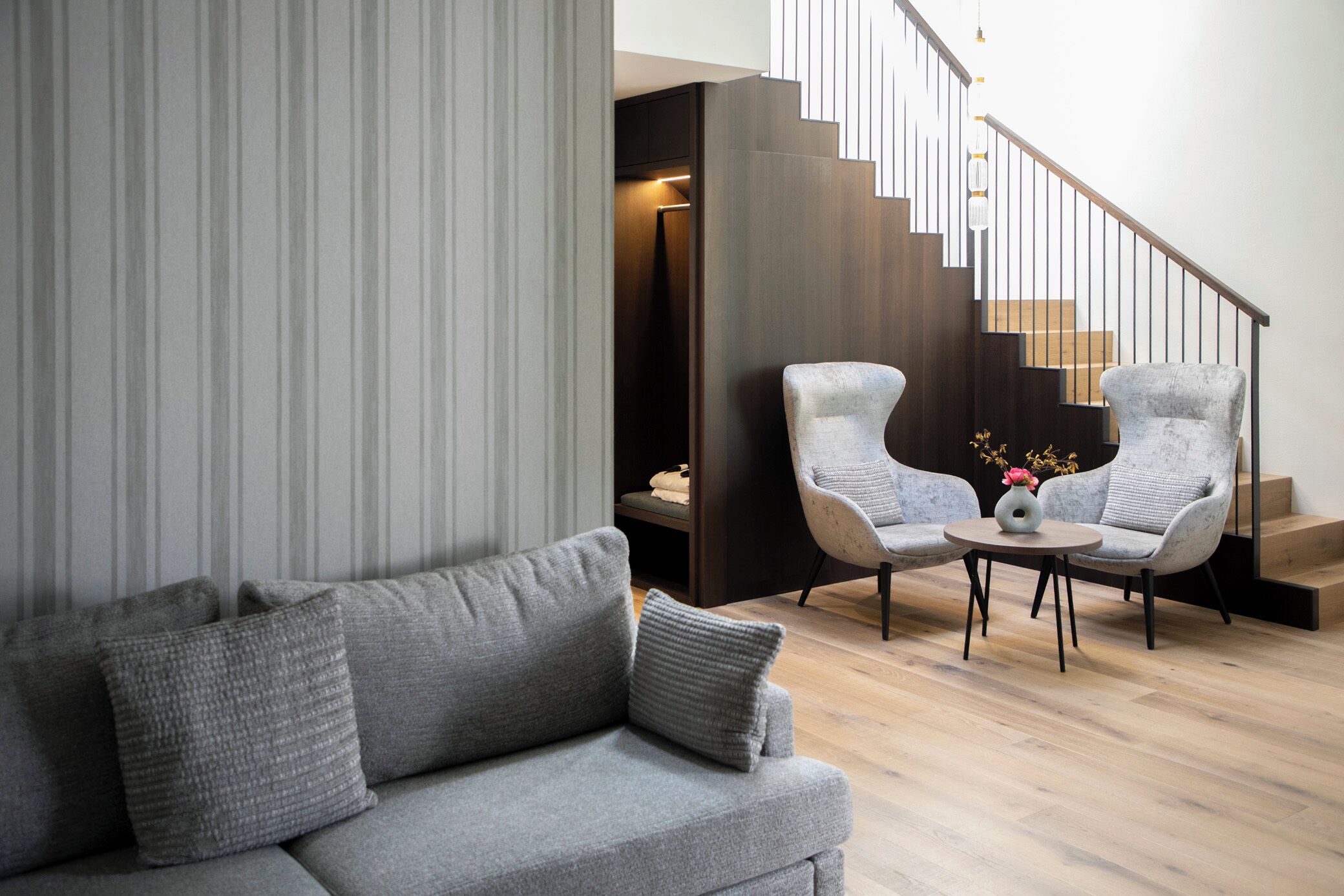 A modern living room with a grey sofa, two armchairs, a coffee table, and a staircase with wooden steps and a metal railing.