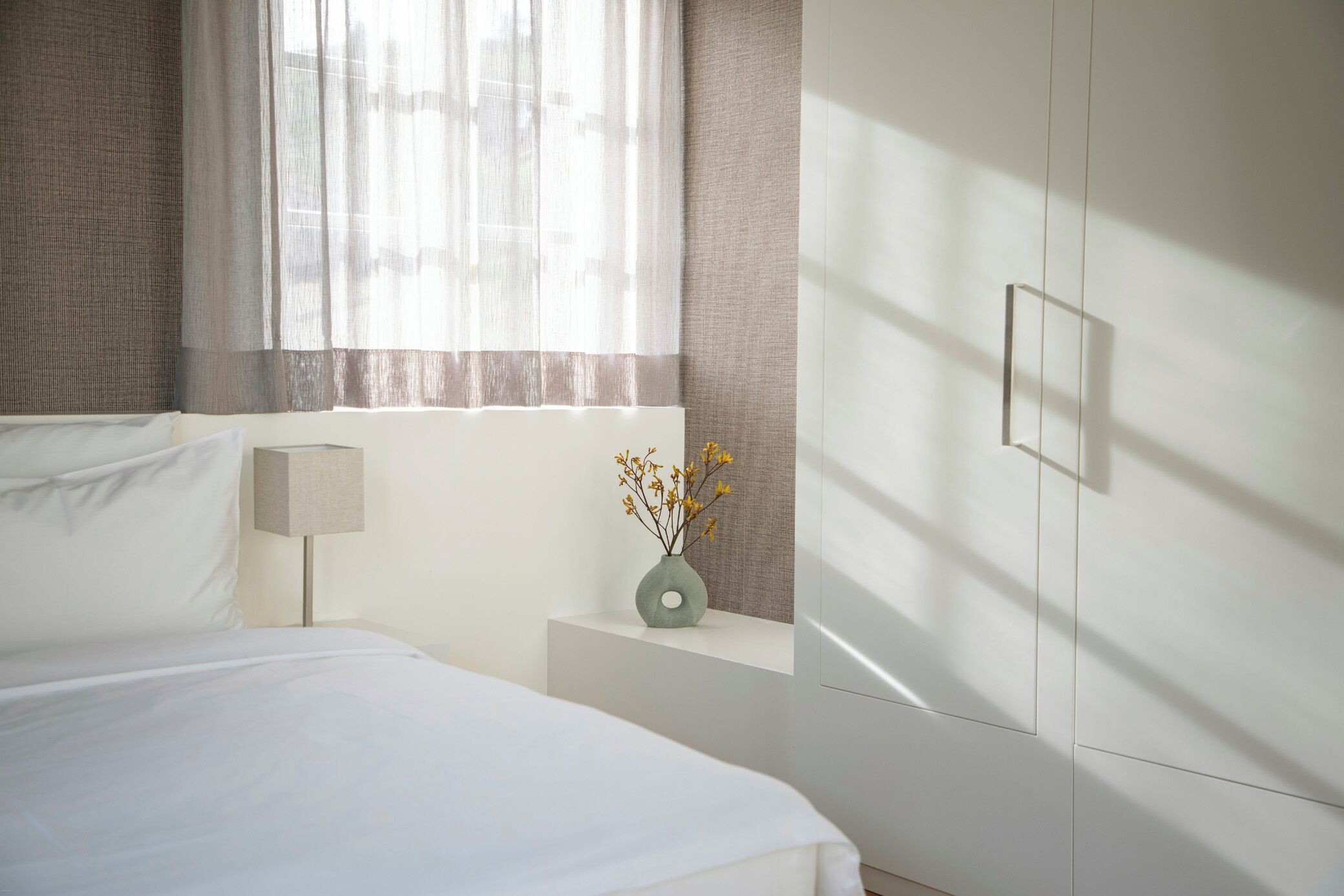 Une chambre à coucher avec un lit, une table de chevet, une lampe, un vase de fleurs et une fenêtre avec des rideaux légers.