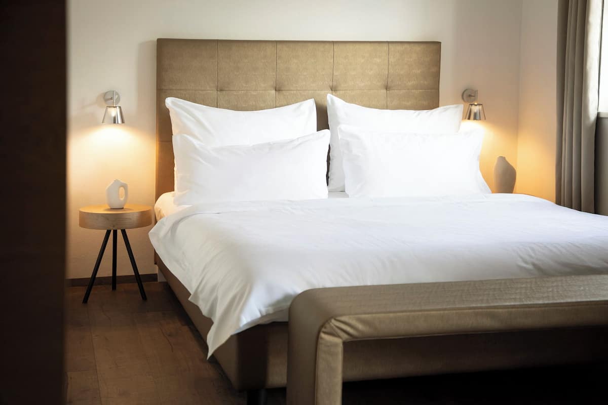 Una cama bien hecha con cabecera capitonada, dos lámparas de noche y una mesita redonda con un cuenco.