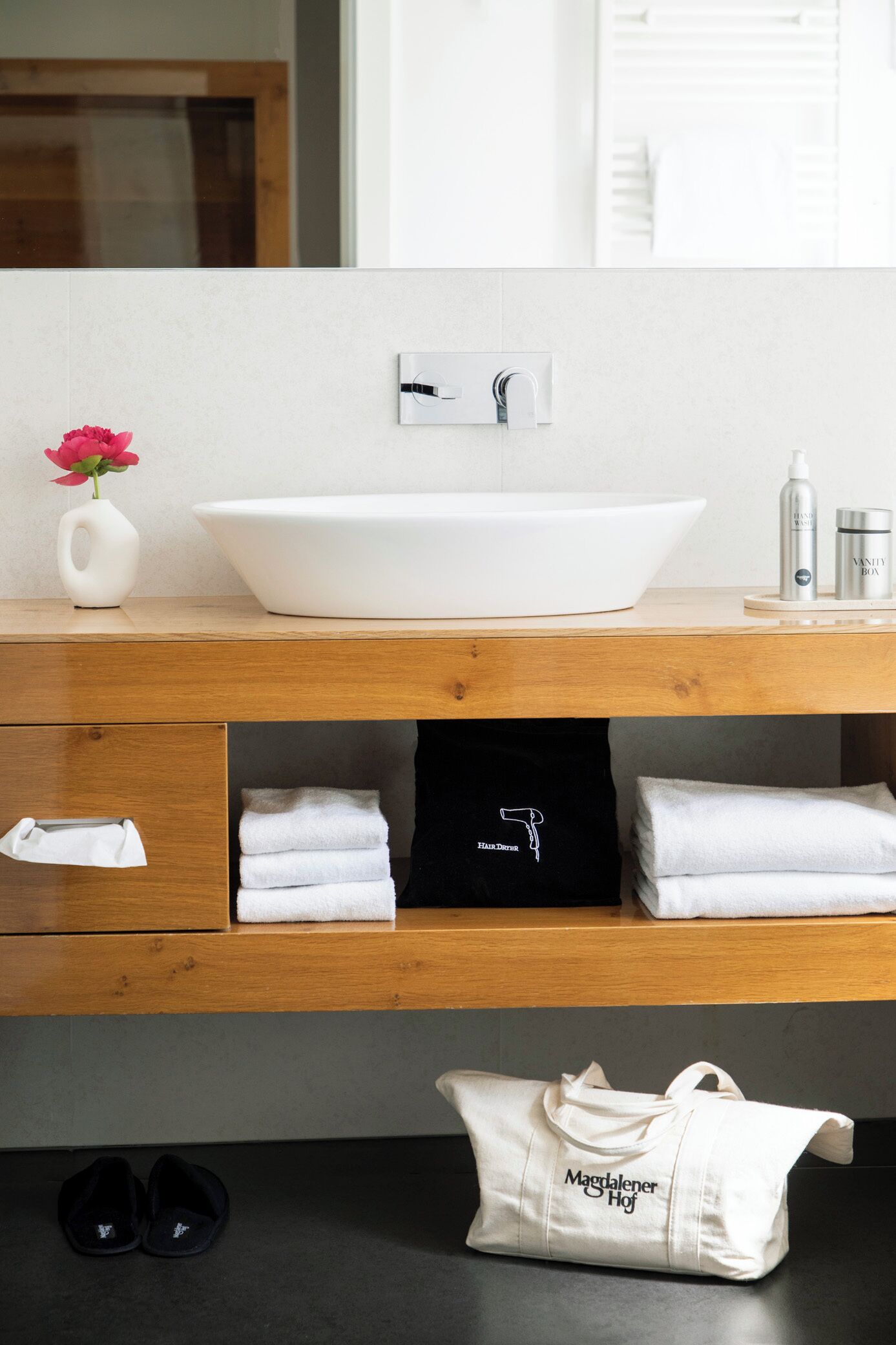 A bathroom with a white vessel sink, wooden vanity, towels, a black bag, and a pair of slippers.