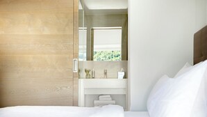 A modern hotel room with a wooden wall, a white bed, a white vanity with a mirror, and a window with a view of greenery. - Magdalenerhof (Bolzano)