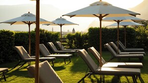 Outdoor lounge chairs with umbrellas, a grassy area, and a distant view of a cityscape with a prominent building. - Magdalenerhof (Bolzano)