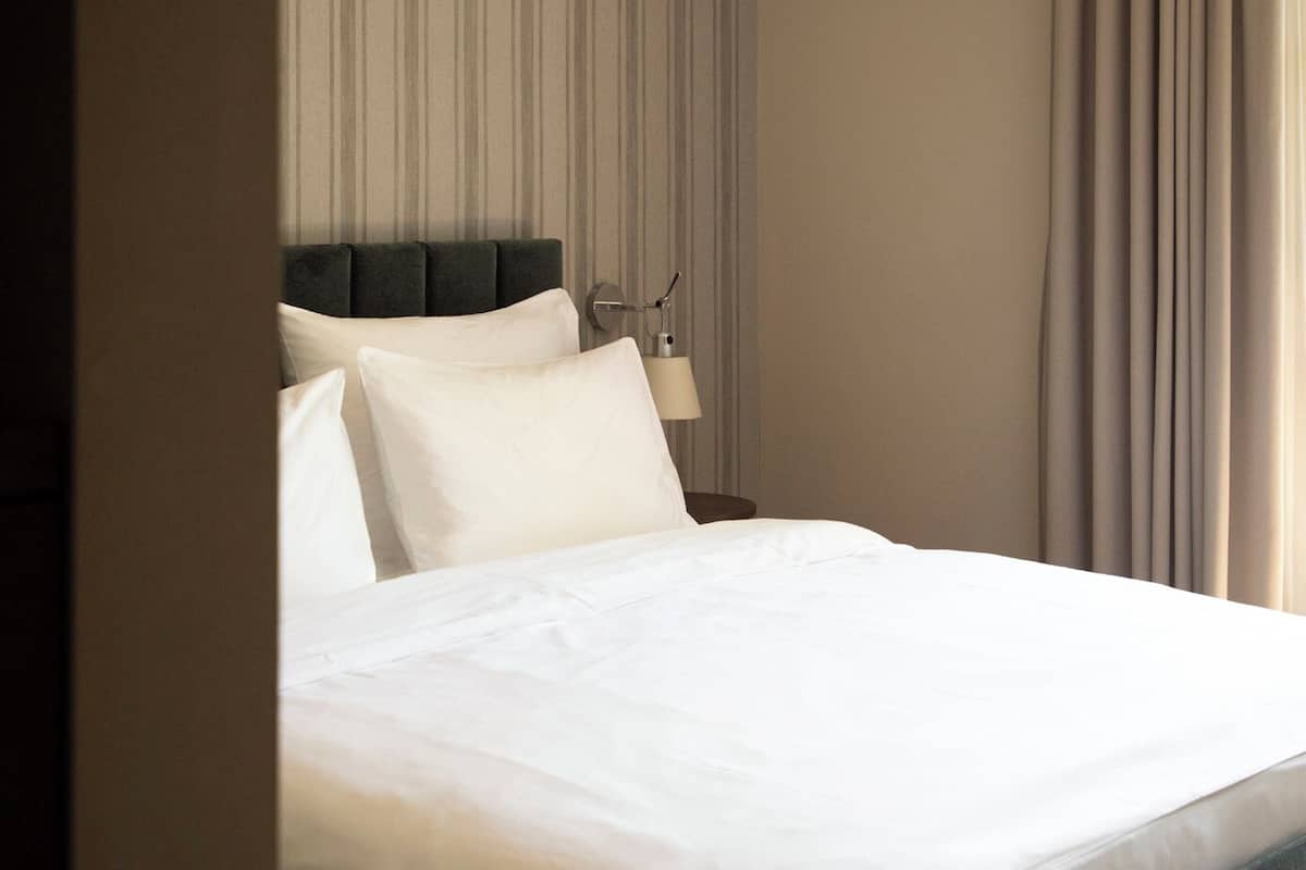 Una habitación de hotel moderna con una cama grande, cabecera, mesita de noche con lámpara y vistas al exterior a través de cortinas.