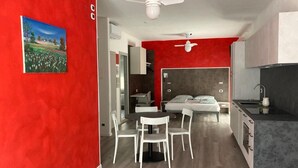 Een moderne kamer met een rode accentmuur, een bed met een grijze hoofdbord, een witte eettafel met stoelen, en een keuken met een grijs aanrecht.