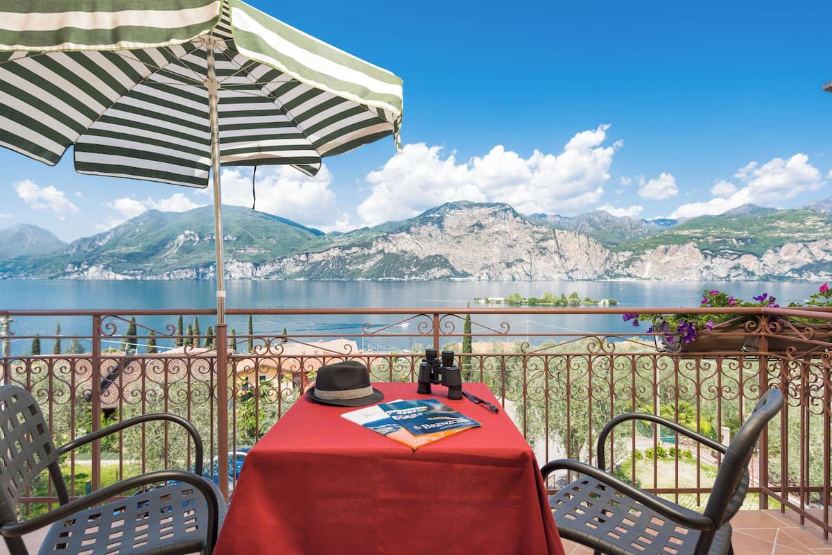 Una mesa con mantel rojo, un paraguas a rayas y un sombrero, con vistas a un lago y montañas al fondo.