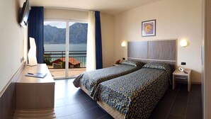 A hotel room with a bed, a desk, a chair, a phone, and a view of the sea and mountains. - Hotel Anna (Malcesine)
