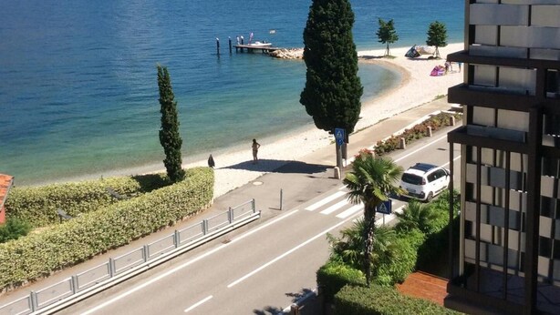 A view of a lake with a dock, a road with a pedestrian crossing, and a building with a modern design. - Hotel Anna (Malcesine)