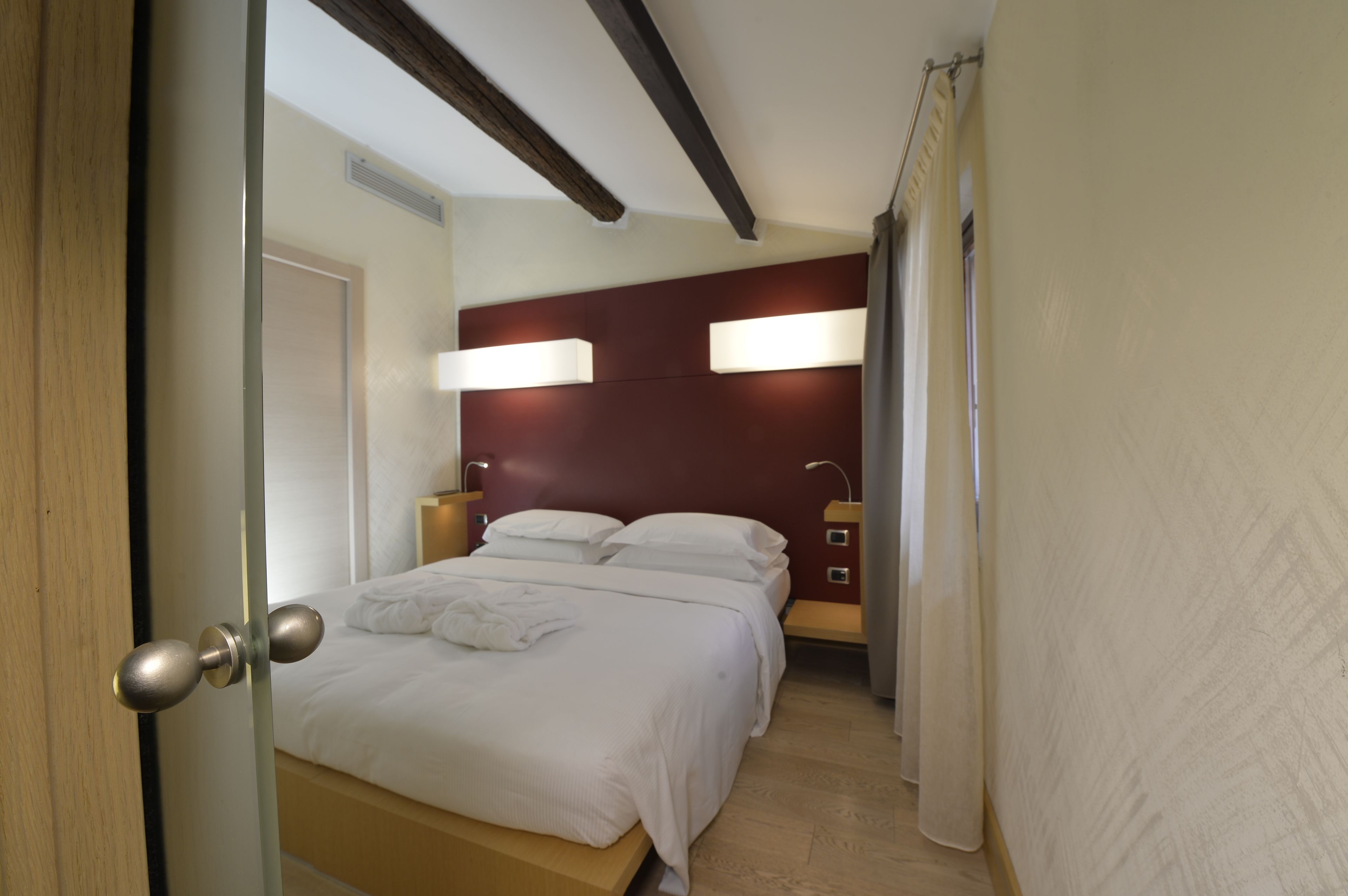 A hotel room with a wooden ceiling, a bed with white linens, two bedside lamps, and a curtain on the window.