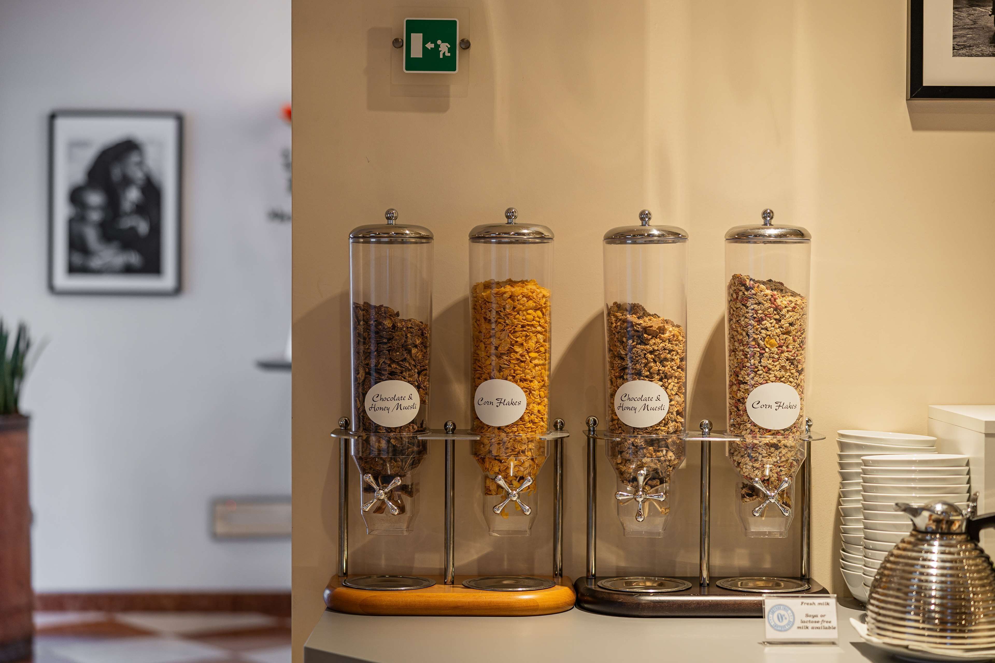 Zona de buffet con cuatro dispensadores de vidrio etiquetados como “Chocolate y Muffin de Miel” y “Corn Flakes”.