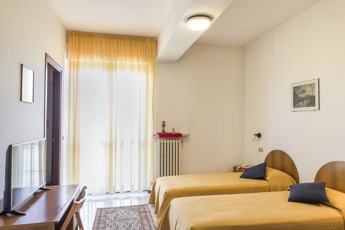 A hotel room with two beds, a desk with a TV, a radiator, and a window with curtains.