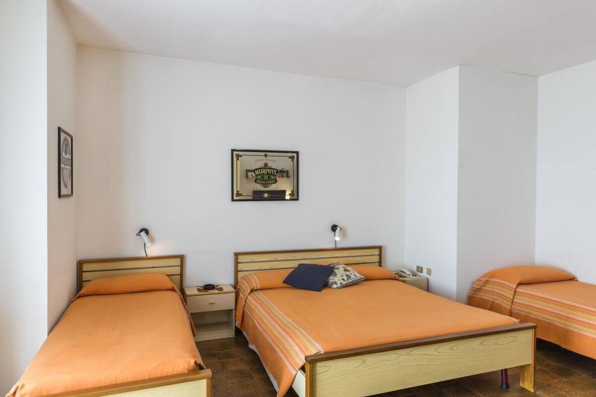 A hotel room with two single beds, each with an orange bedspread and a wooden headboard. There is a framed picture on the wall above the beds, and a small bedside table with a lamp on each bed.