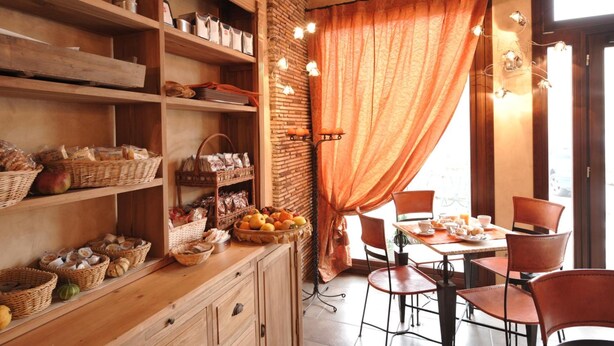 A rustic cafe interior with wooden shelves stocked with bread and pastries, a table set for two with chairs, and a large window with orange curtains.