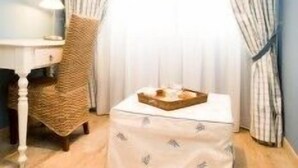 A room with a wooden floor, a white table with a lamp, a woven chair, and a small ottoman with a tray.