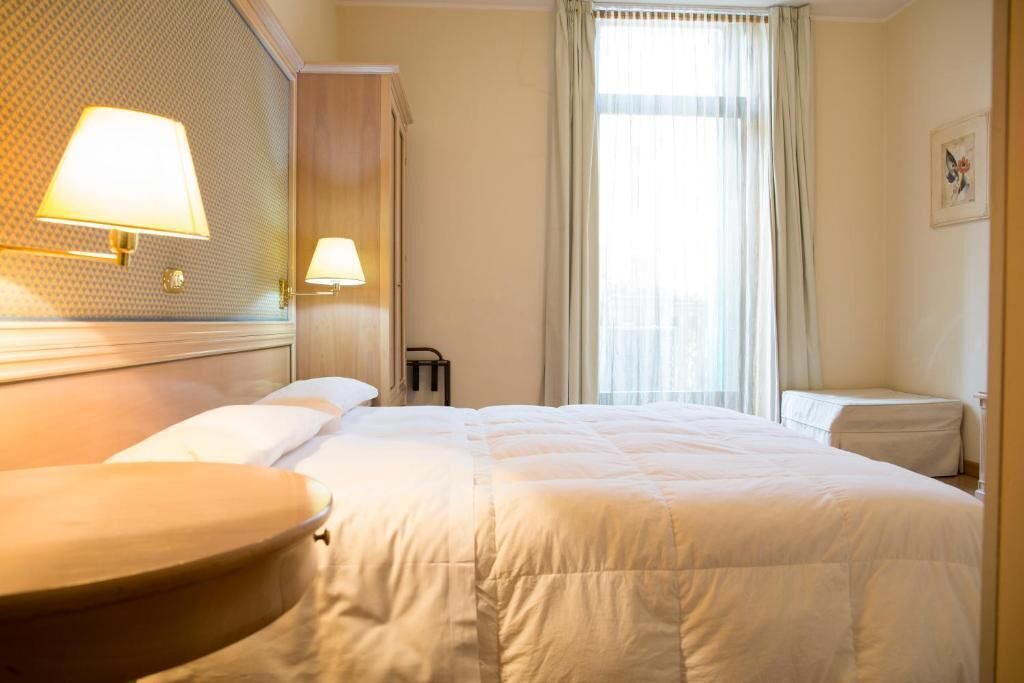 A hotel room with a large bed, two bedside lamps, a window with curtains, and a wall-mounted light fixture.