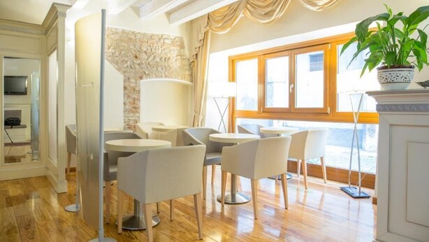A modern living room with a stone wall, wooden flooring, and a large window with curtains. There are several white chairs and a small table. - Hotel al Castello (Verona)