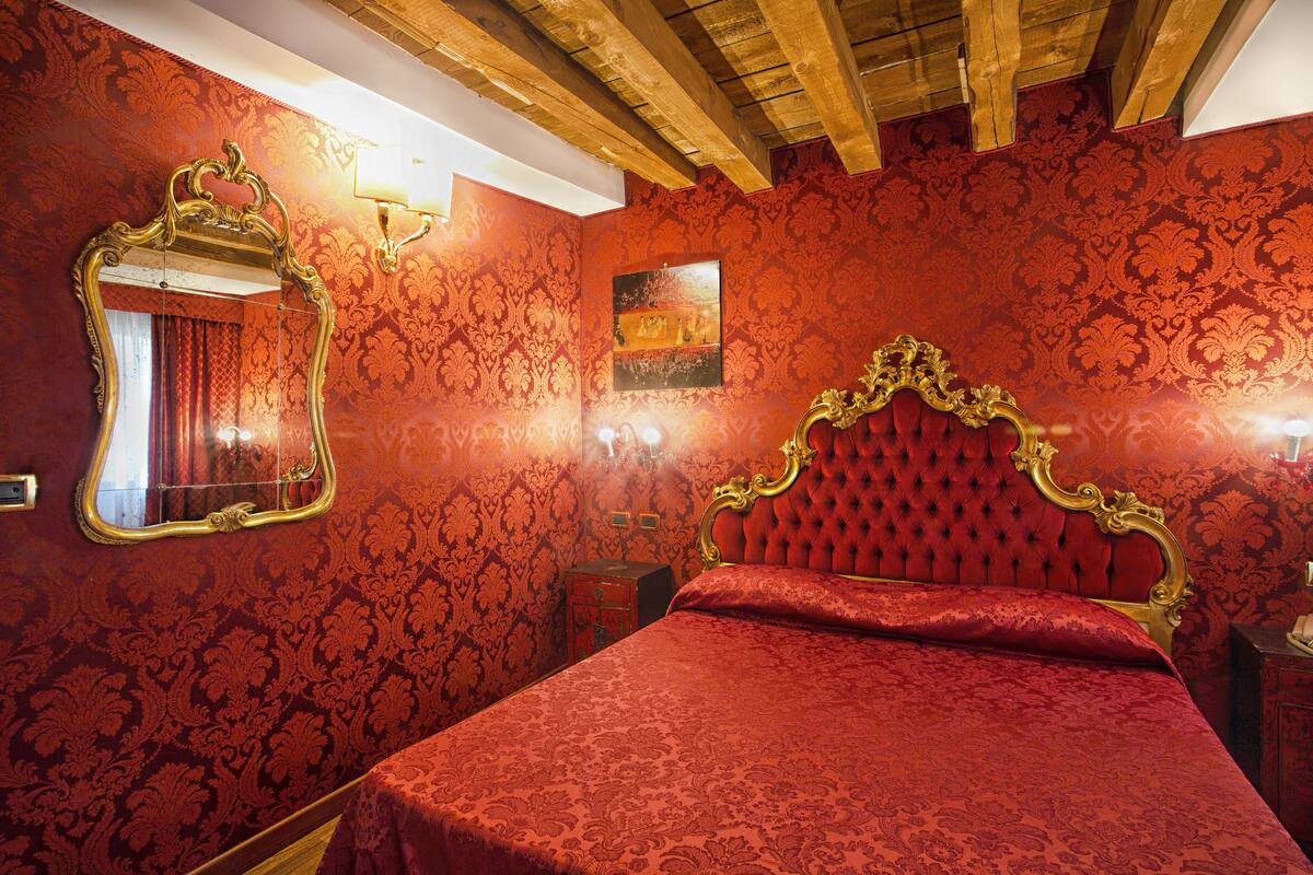 A room with a red patterned wallpaper, a large ornate mirror, a tufted headboard, and a wooden ceiling.