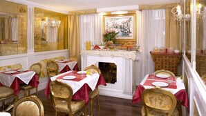 A dining area with tables set for four, gold chairs, a fireplace, and a painting above the mantel. - Hotel Noemi (Venice)