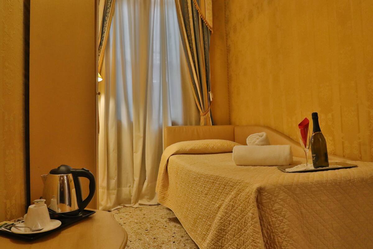 A hotel room with a bed, a bedside table with a kettle, a wine bottle, and a glass.