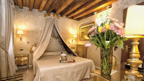 A bedroom with a four-poster bed, a bedside table with a lamp, a vase of flowers, and a mirror. - Hotel Noemi (Venice)