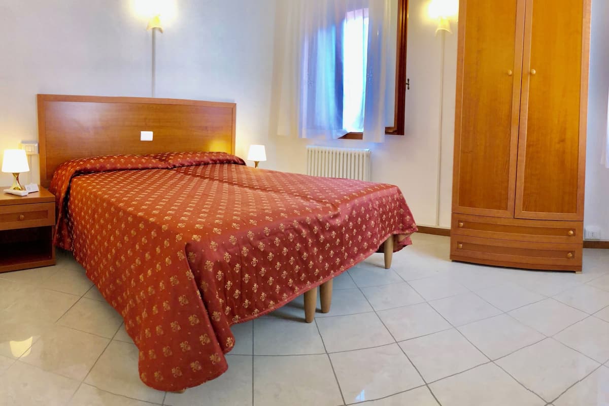 A hotel room with a large bed, two bedside tables with lamps, a wooden wardrobe, and a radiator.