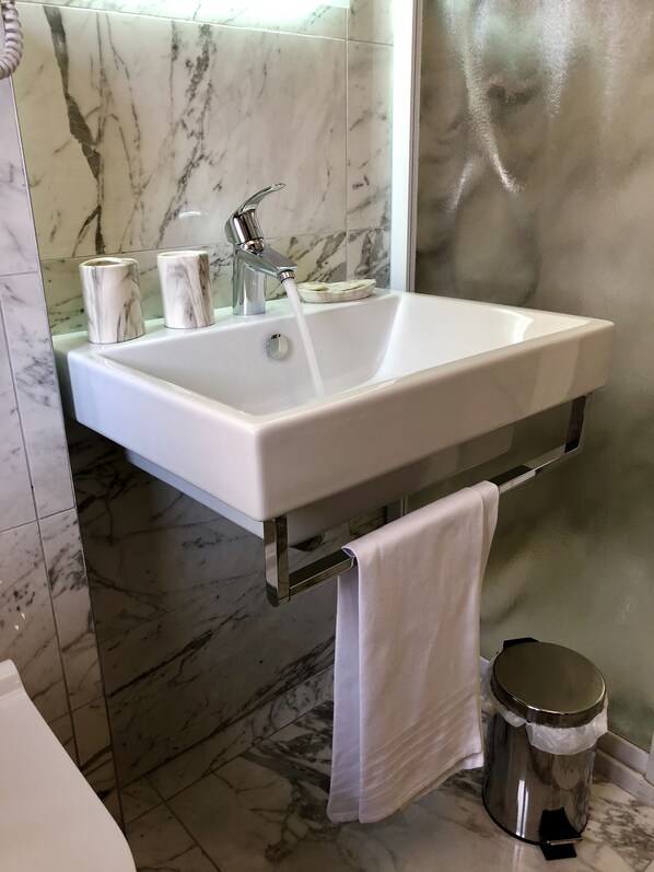 A bathroom with a white sink, marble tiles, a towel rack, and a trash bin.