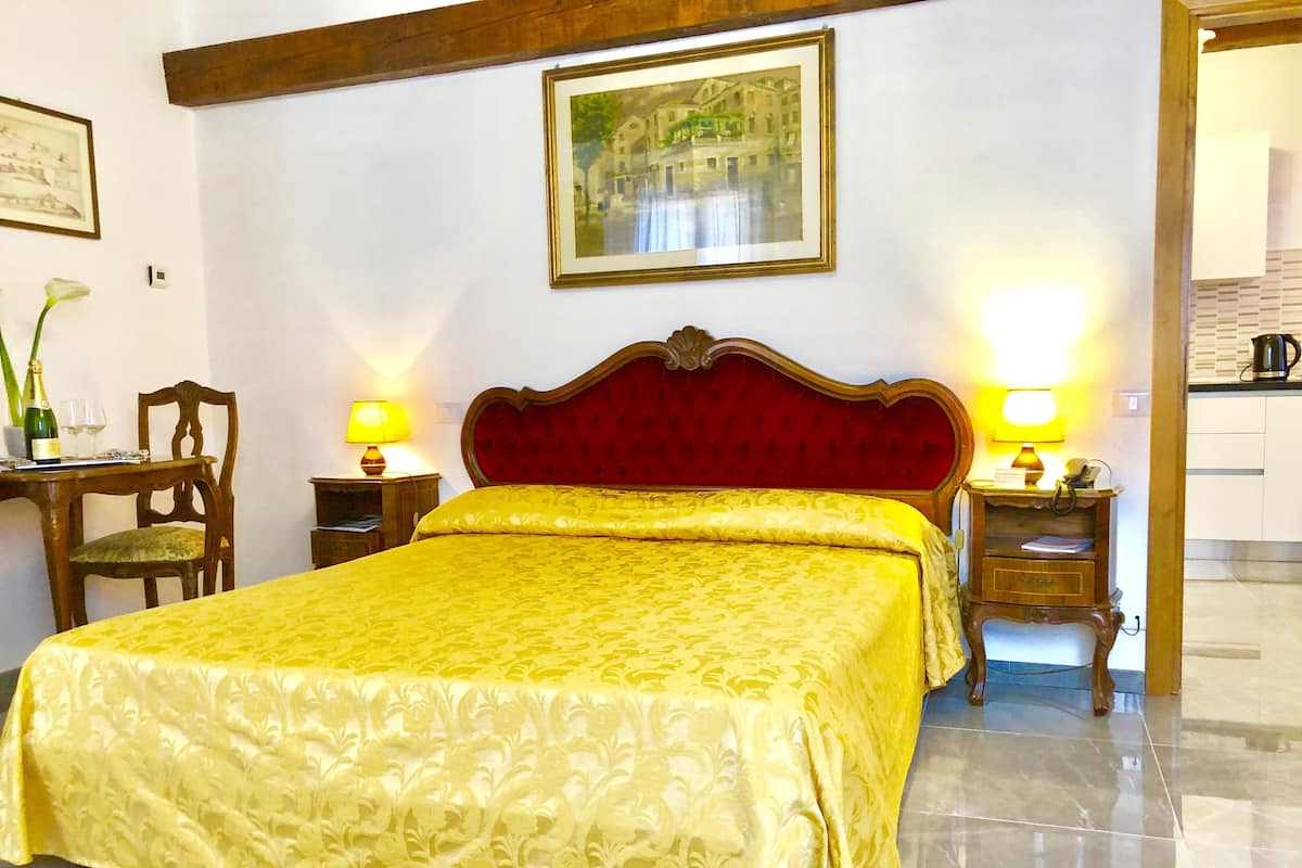 A hotel room with a large bed, two bedside tables with lamps, a wooden chair, a desk with a chair, a framed picture on the wall, and a kitchen area visible through an open door.