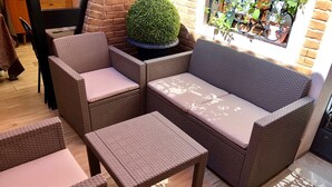 Un canapé en osier avec des coussins, une table ronde surmontée d’un arbuste taillé en forme, et une fenêtre agrémentée de plantes en pot.