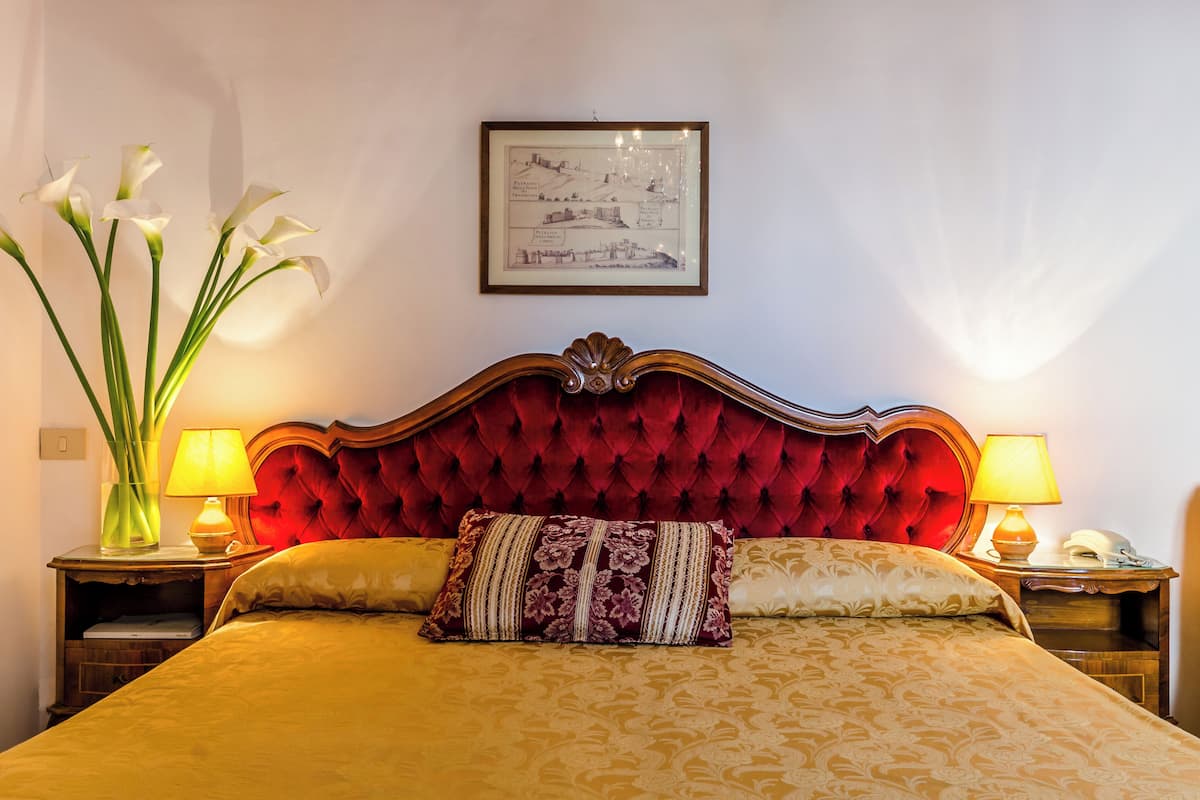 A bed with a red headboard, two bedside lamps, a vase with flowers, and a framed picture on the wall.