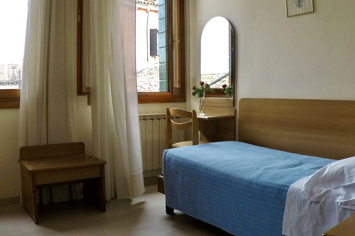 Un dormitorio con una cama individual, un banco de madera, una mesita con un florero y una ventana con cortinas.