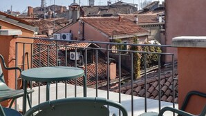 Una terraza con sillas de metal y una mesa, con vistas a un paisaje urbano con edificios tradicionales y el campanario de una iglesia.