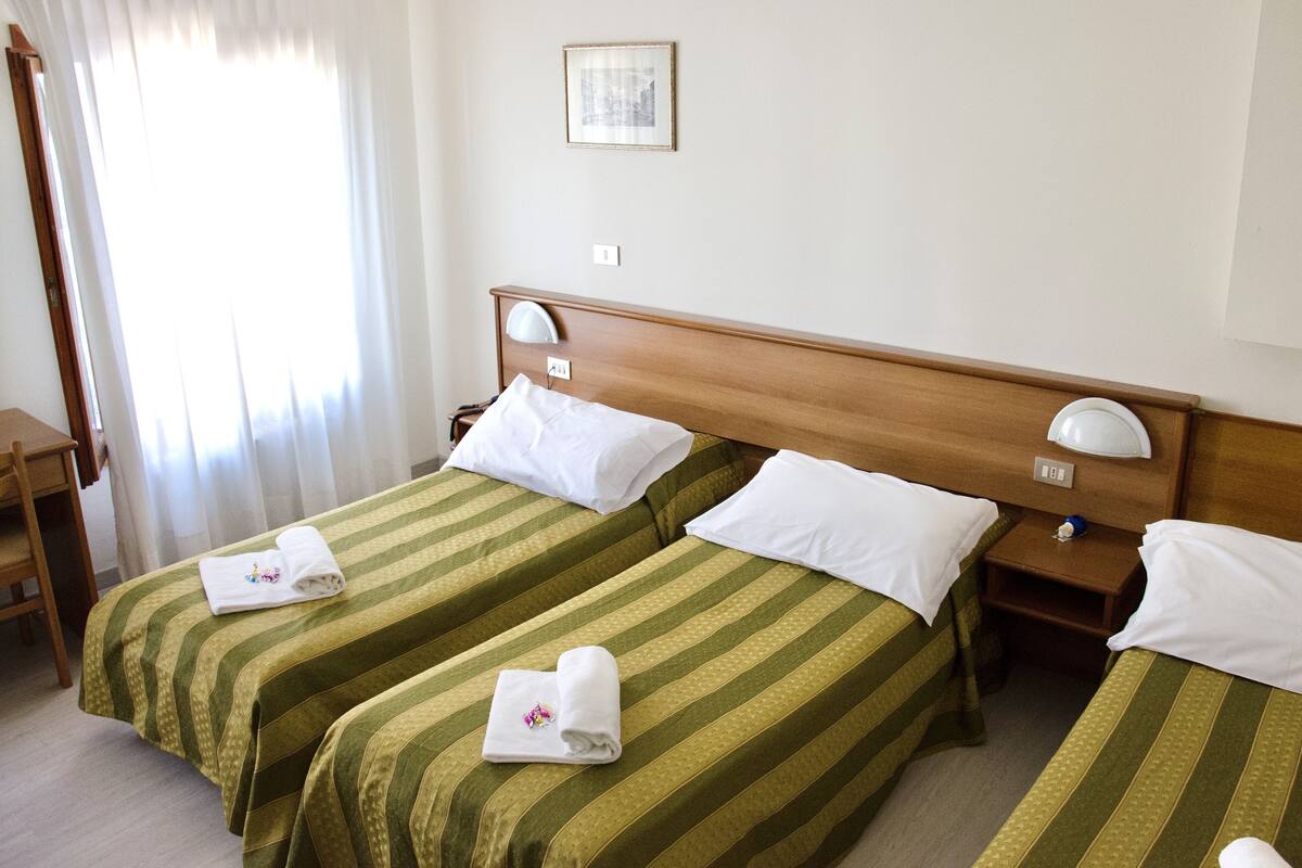 Habitación de hotel con dos camas individuales, cada una con una colcha verde a rayas y almohadas blancas. Tiene un cabecero de madera, una mesita de noche con una lámpara y un cuadro enmarcado en la pared.