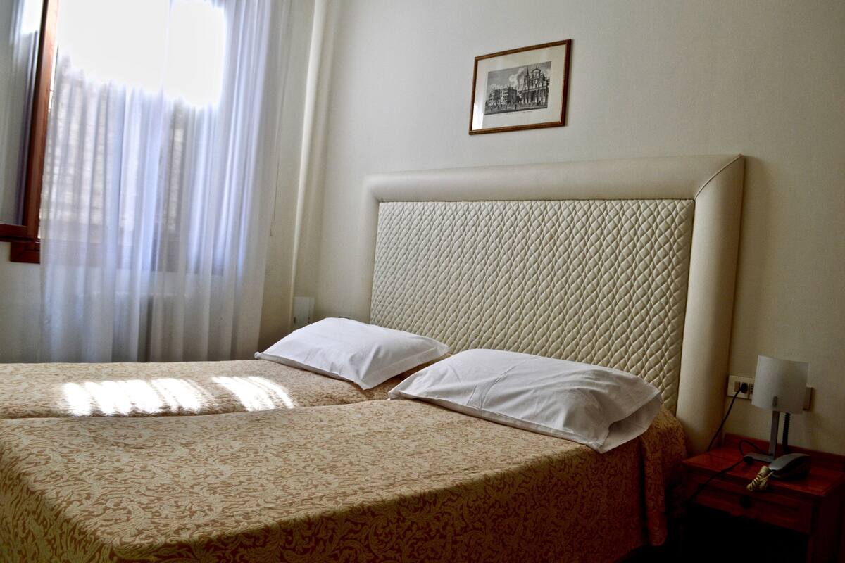 Una habitación de hotel con dos camas, un cabecero acolchado, una mesita de noche con una lámpara y un cuadro enmarcado en la pared.
