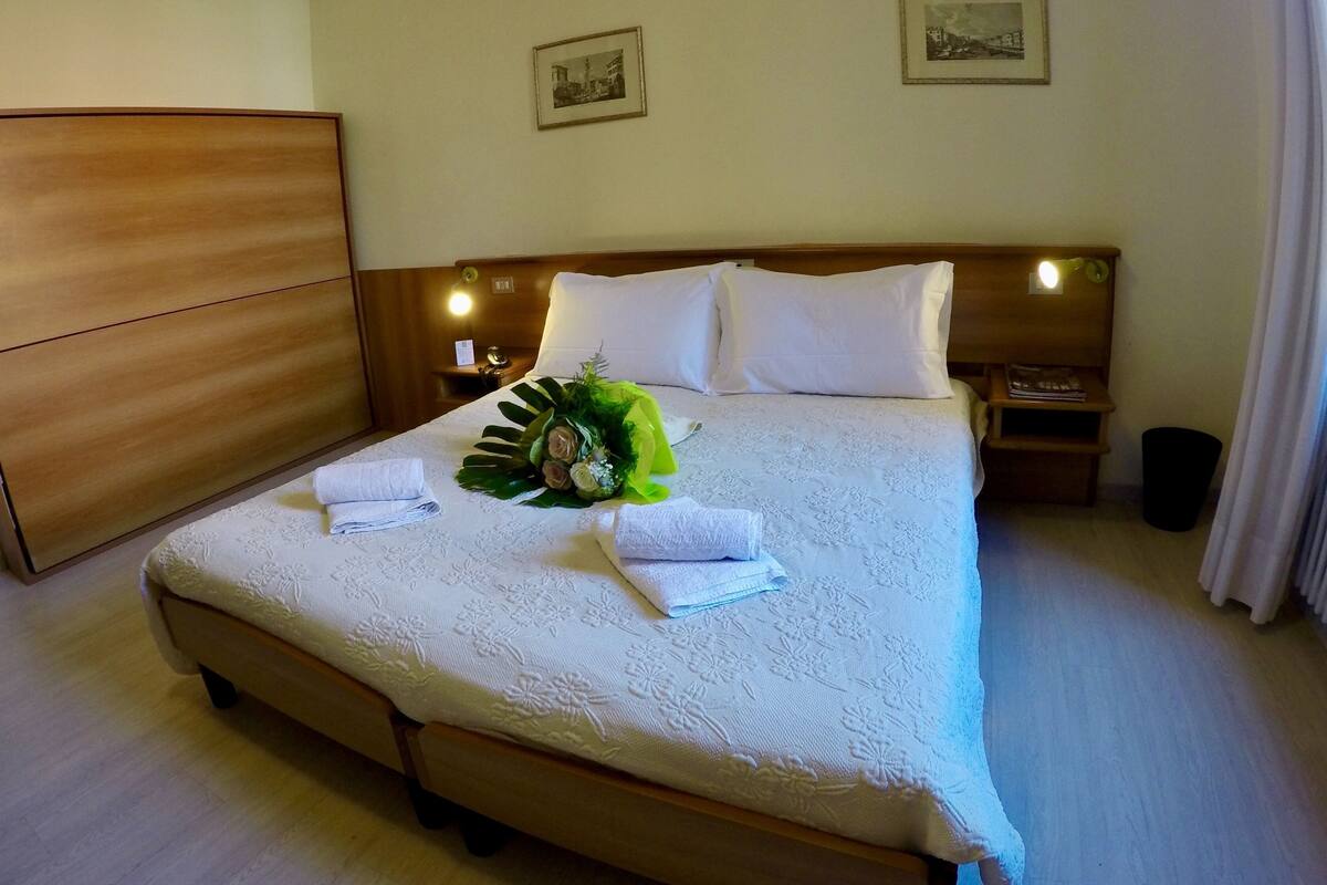 Una cama bien hecha con sábanas blancas, un edredón estampado con motivos florales y un ramo de flores. Hay dos mesitas de noche con lámparas, un armario de madera y dos cuadros en la pared.