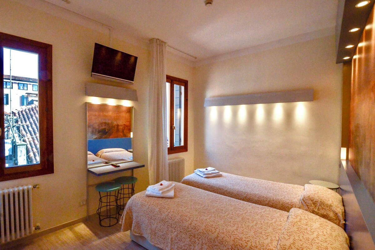 Habitación de hotel con dos camas, un escritorio con silla, un espejo y ventana con vista a edificios.